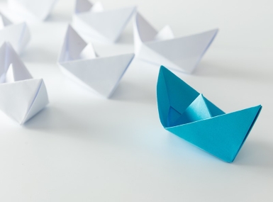 A blue paper boat leads ahead of rows of white paper boats